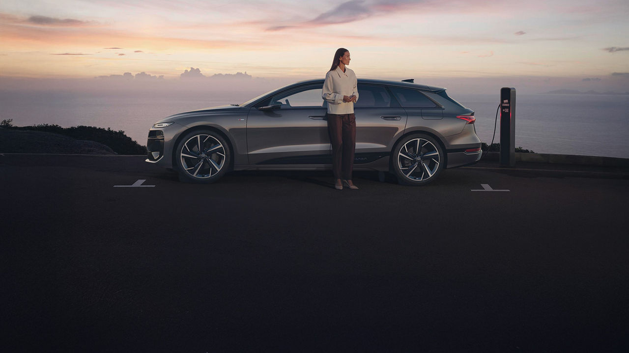 Frau steht neben einem silbernen Audi an einer Ladesäule mit Meerblick bei Sonnenuntergang.